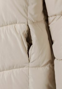 Sezione di giacca a vento beige con texture a quadrati, dotata di apertura per tasca laterale. Tessuto morbido e liscio con una finitura leggermente lucida.