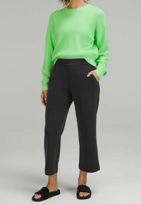 Person som bär en neongrön tröja, svarta cropped byxor och svarta slide-sandaler står med en hand i fickan mot en grå bakgrund.