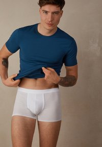 Blått figursytt kortärmat t-shirt med en slät textur, bärs ovanpå vita boxertroser. Modellen har en synlig tatuering på en arm.