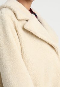 Persona che indossa un cappotto di shearling oversized di colore crema, con una texture, e un ampio colletto sopra una camicia a quadri rossi.