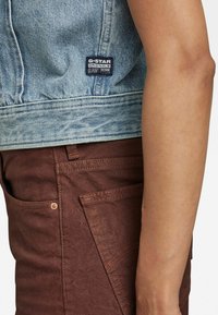 Ljusblå denimjacka med en G-Star-lapp, i kombination med bruna texturerade byxor med synlig sömnad och sidofickor.