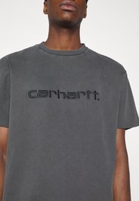 Grijze katoenen t-shirt met een ronde hals, voorzien van een contrasterend zwart geborduurd "carhartt" logo op de borst. Korte mouwen.