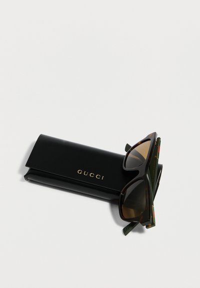 Lunettes de soleil Gucci noires avec monture écaille de tortue reposant sur un étui en cuir noir Gucci avec lettrage doré sur fond blanc.