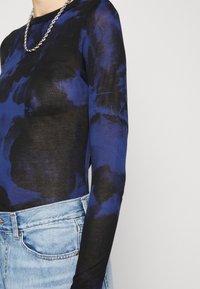 Maglietta a maniche lunghe in tessuto nero trasparente con motivo tie-dye blu. Caratterizzata da un collo rotondo e un piccolo logo su un lato. Abbinata a jeans azzurri chiari.