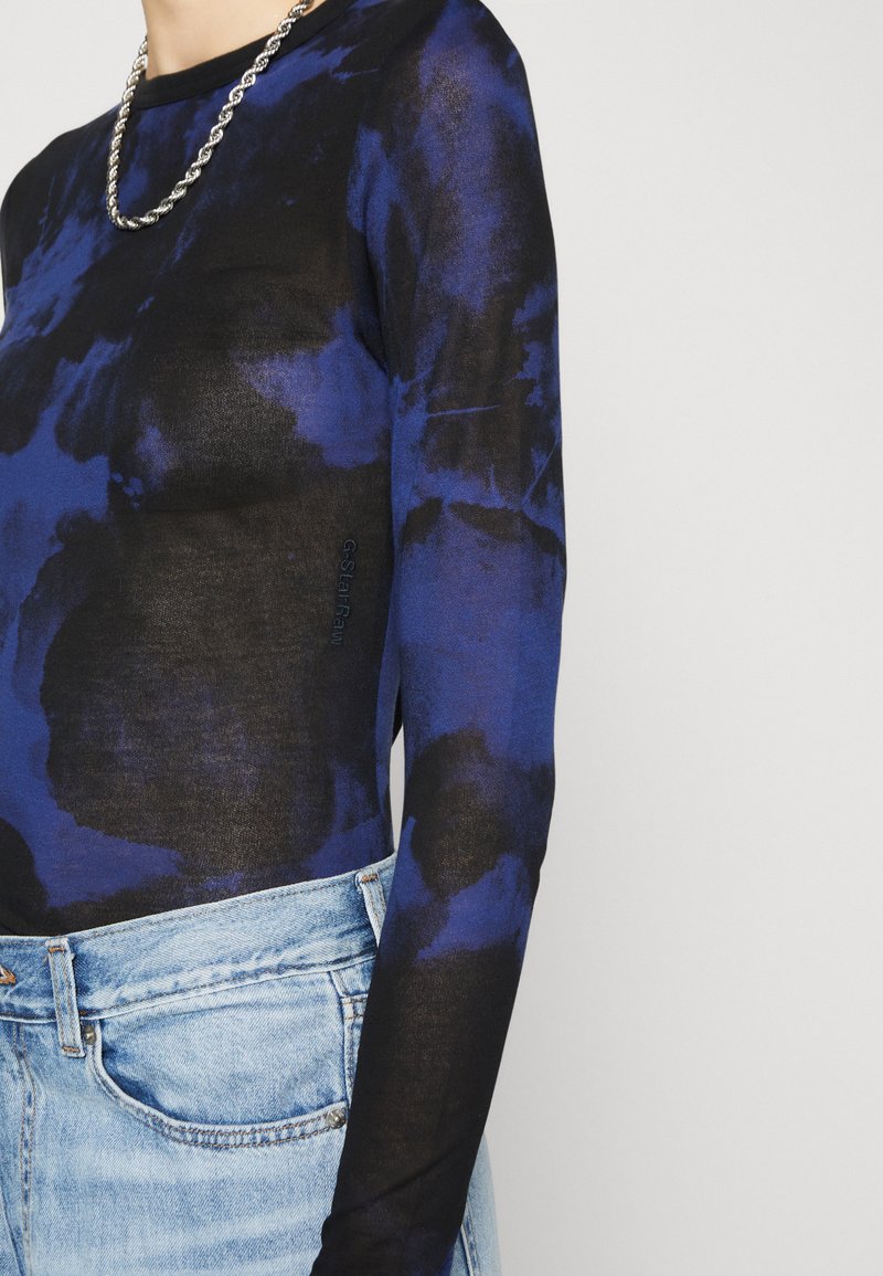 Długi rękaw bluzka z przezroczystej czarnej tkaniny z niebieskim wzorem tie-dye. Posiada okrągły dekolt i małe logo po jednej stronie. Połączona z jasnoniebieskimi dżinsami.