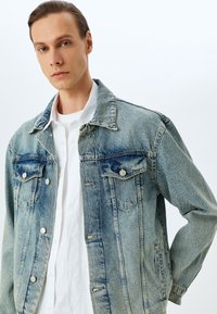 Giacca di jeans, azzurra chiara, leggermente sbiadita, con chiusura a bottoni, due tasche sul petto e colletto a punta, indossata sopra una camicia bianca.