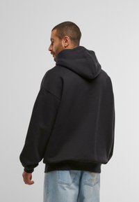 Schwarzer Hoodie mit großer Rückentasche, oversized Passform und Kapuze mit Kordelzug. Getragen mit ausgewaschenen blauen Jeans mit Gesäßtaschen.