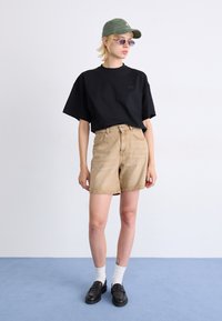 T-shirt nera a maniche corte, pantaloni corti in denim khaki, cappellino verde, occhiali da sole, calzini bianchi e mocassini neri su uno sfondo chiaro.