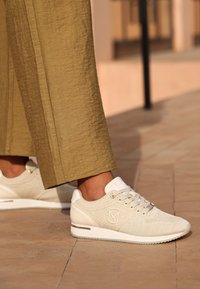 Beiga stickade sneakers med snörningsdesign, vita accenter och logodetalj på sidan, med en strukturerad yta och solid gummisåle.