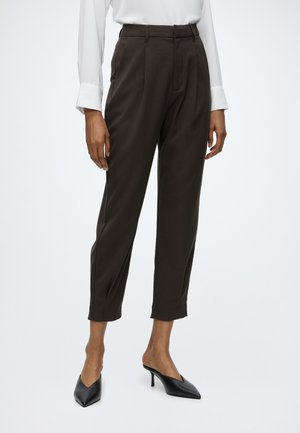 Personne portant un pantalon marron foncé sur mesure longueur cheville, une chemise blanche à manches longues et des talons hauts noirs pointus.