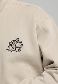 Beige sweatshirt featuring a black embroidered figure in motion, set against a textured fabric background with visible stitching details.