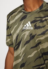 Camiseta de Adidas con patrón de camuflaje verde, de mangas cortas, fabricada en tejido transpirable, con un logo blanco en la parte superior izquierda del pecho.