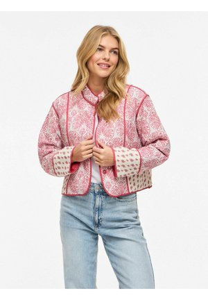 Smilende kvinde med langt blondt hår iført en lyserød og hvid mønstret quiltet jakke over en hvid top og lyseblå jeans.