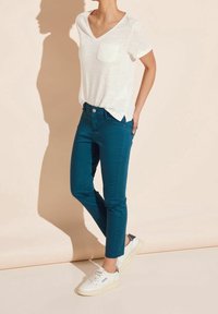 Bílé tričko s krátkým rukávem a výstřihem do V s kapesníčkem na hrudi, doplněné o tyrkysové úzké kalhoty a bílé tenisky, na neutrálním pozadí.