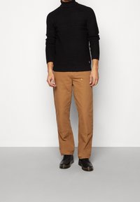 Pull noir côtelé à col roulé avec manches longues, associé à un pantalon beige avec poches avant et coupe décontractée, complété par des bottes noires.