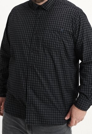 Mann mit einem schwarz-grau karierten Hemd mit langen Ärmeln und einer Brusttasche vorne, kombiniert mit grauer Hose.