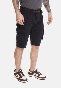 Pantaloni cargo neri con molte tasche laterali, realizzati in materiale resistente, dotati di cintura e dal taglio rilassato. Indossati con scarpe da ginnastica marroni e bianche.