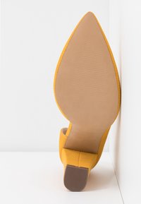 Chaussure à talon haut jaune avec un bout pointu, un extérieur texturé et une semelle beige. Le talon est fin et effilé, offrant un profil élégant.