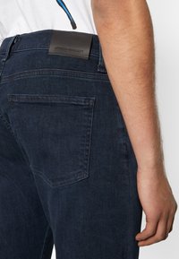 Mörkblå denimjeans med en läderetikett på midjebandet. Har raka ben och en bakficka. Minimalistisk design.