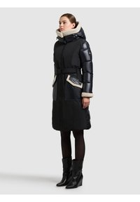 Lange zwarte pufferjas met een ceintuur in de taille, een zachte capuchonrand en contrasterende zakaccenten. Draagt zwart sleehakkenlaarzen en donkere panty's.