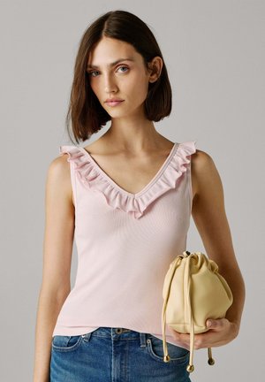 Débardeur rose clair avec col en V et bordure à volants, associé à un sac beige arrondi avec cordon de serrage. Le mannequin porte un jean en denim.