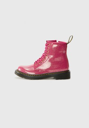 Skinnende pink glitrende snørestøvle med sort sål og gule syninger, vist i sideprofil på en ensfarvet baggrund.