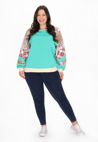 Felpa turchese con maniche decorate, caratterizzate da motivi a quadri e floreali. Abbinata a jeans skinny blu scuro e sneakers bianche.