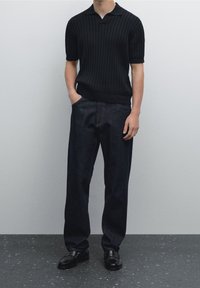 Czarna prążkowana koszulka polo z krótkim rękawem, zestawiona z luźnymi ciemnymi dżinsami. Model nosi czarne skórzane mokasyny, ma ręce w kieszeniach.