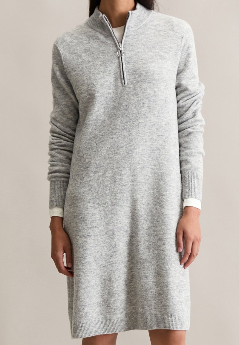Femme portant une longue robe-pull grise en maille avec col à demi-fermeture éclair et poignets côtelés, debout devant un fond uni.