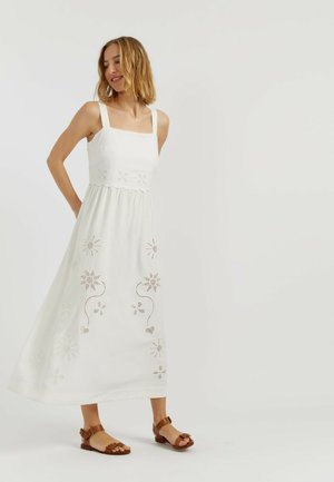 Femme portant une robe midi blanche sans manches avec des découpes florales et des sandales plates marron, debout devant un fond clair uni.