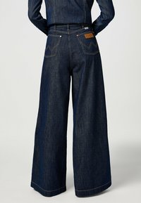 Jeans in denim a gamba larga di colore blu scuro, con vita alta, due tasche posteriori con cuciture a zigzag e cuciture arancioni a contrasto sui orli.