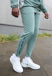 Světlé zelené sportovní kalhoty s úzkým střihem, zdobené textem "Žádný člověk není omezen", spárované s bílými sportovními botami.