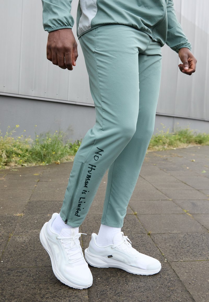 Světlé zelené sportovní kalhoty s úzkým střihem, zdobené textem "Žádný člověk není omezen", spárované s bílými sportovními botami.