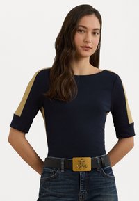 Haut bleu marine avec des accents dorés aux épaules, manches courtes et col rond. Porté avec un jean en denim foncé et une ceinture noire avec une boucle en or.