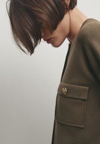 Olive green knit cardigan with a textured finish, featuring a front pocket and a gold decorative button.  The collar is rounded.