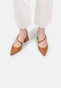 Picioare purtând pantofi cu toc bloc ascuțit din piele bronzată, cu curea diagonală, asociați cu pantaloni albi scurți, pe un fundal alb.