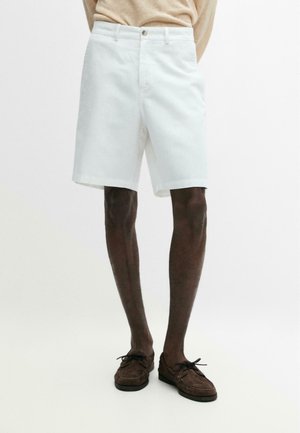 Uomo con pantaloncini bianchi fino al ginocchio e scarpe marroni con lacci, in piedi contro uno sfondo bianco neutro.