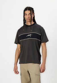 Dark gray t-shirt with a round neckline, featuring a horizontal navy stripe and white accent line, and a logo in the center. Short sleeves.