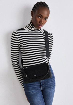 Jeune femme aux cheveux tressés portant un col roulé rayé noir et blanc, un jean bleu et un sac bandoulière noir, debout contre un mur blanc.