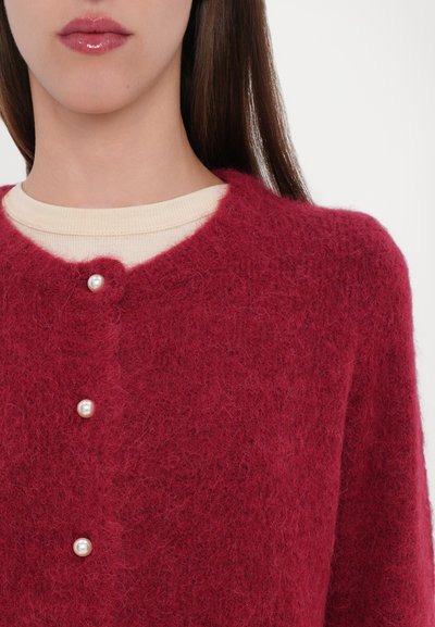 Cardigan rosso porpora in peluche con scollo rotondo, caratterizzato da tre bottoni perla davanti, indossato sopra una camicia di colore crema.