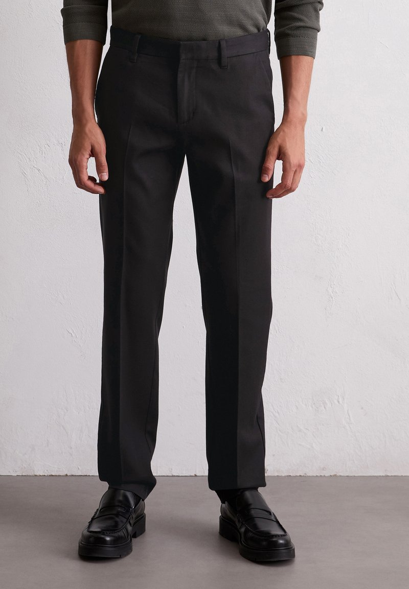 Pantaloni neri sartoriali con taglio dritto, dotati di passanti per cintura e una superficie liscia, abbinati a mocassini neri in pelle.