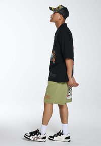 Maglietta nera a maniche corte con stampa grafica, pantaloni corti verde oliva con ricami e sneakers nere con accenti rossi. Vestibilità rilassata.