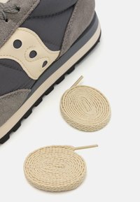 Gray sneaker with suede and nylon materials, featuring cream accents, a layered design, and two rolled beige laces beside it.
