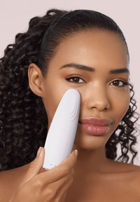 Gezichtsapparaat van wit plastic met een gebogen punt en bedieningsknoppen, tegen de wang gehouden door een persoon met krullend haar en lipgloss.