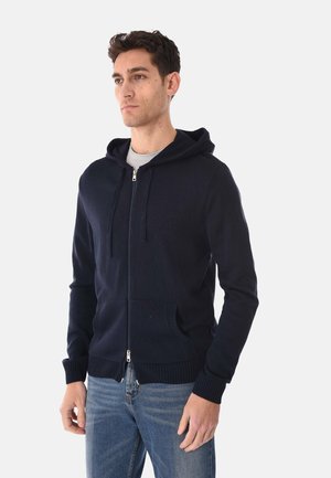 Giovane uomo che indossa una felpa con cappuccio blu navy con cerniera e tasche frontali, e jeans azzurri, in piedi davanti a uno sfondo chiaro e semplice.