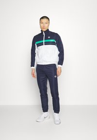 Track suit in navy and white, featuring green stripes across the chest, zip-up jacket, elastic cuffs, and logo on the chest and pant.