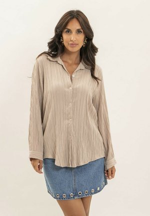 Femme aux longs cheveux foncés portant une chemise beige texturée boutonnée et une jupe en denim bleu avec des détails en œillets métalliques, debout devant un fond uni.