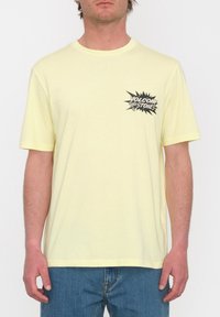 Camiseta de algodón amarilla con un diseño impreso en negro que lleva el texto "VOLCOM" y "THE STONES" en el pecho. Mangas cortas, cuello redondo.