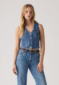 Gilet en denim bleu moyen avec un col en V, des boutons sur le devant et deux poches poitrine. Associé à un jean en denim taille haute et une ceinture noire.
