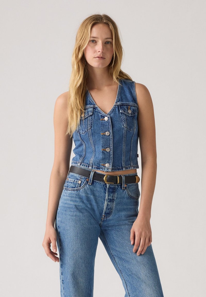 Džinsinis liemenė su V formos iškirpte, priekiniais mygtukais, dviem krūtinės kišenėmis ir trumpintu dizainu, derinamas su aukštos liemens mėlynais džinsais ir juodu diržu.